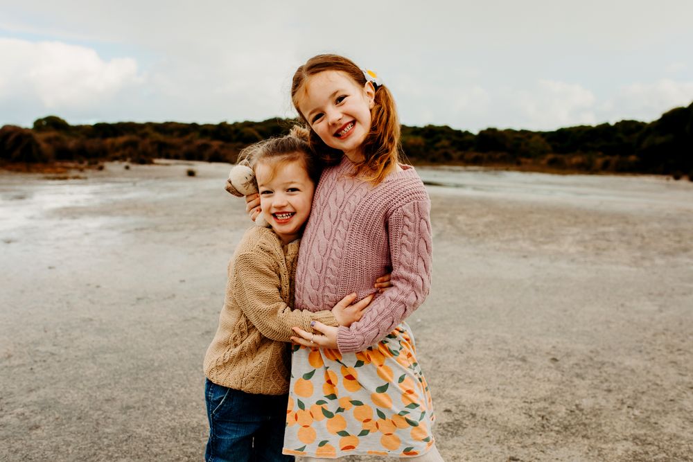 Playful couple laughing and embracing on a sandy beach on an overcast day while wearing casual sweaters.