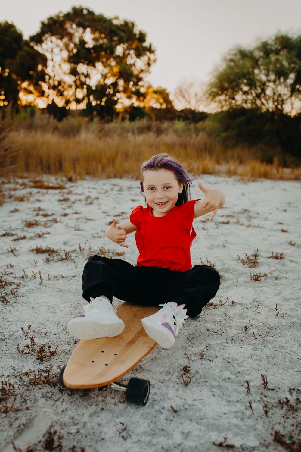 A young skateboarder in a red shirt performs tricks on sandy terrain at sunset.