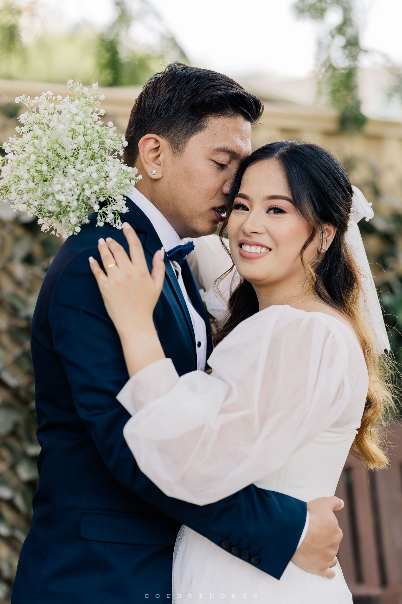 A romantic couple in formal attire sharing tender moments during an outdoor photoshoot with white flowers in the background.