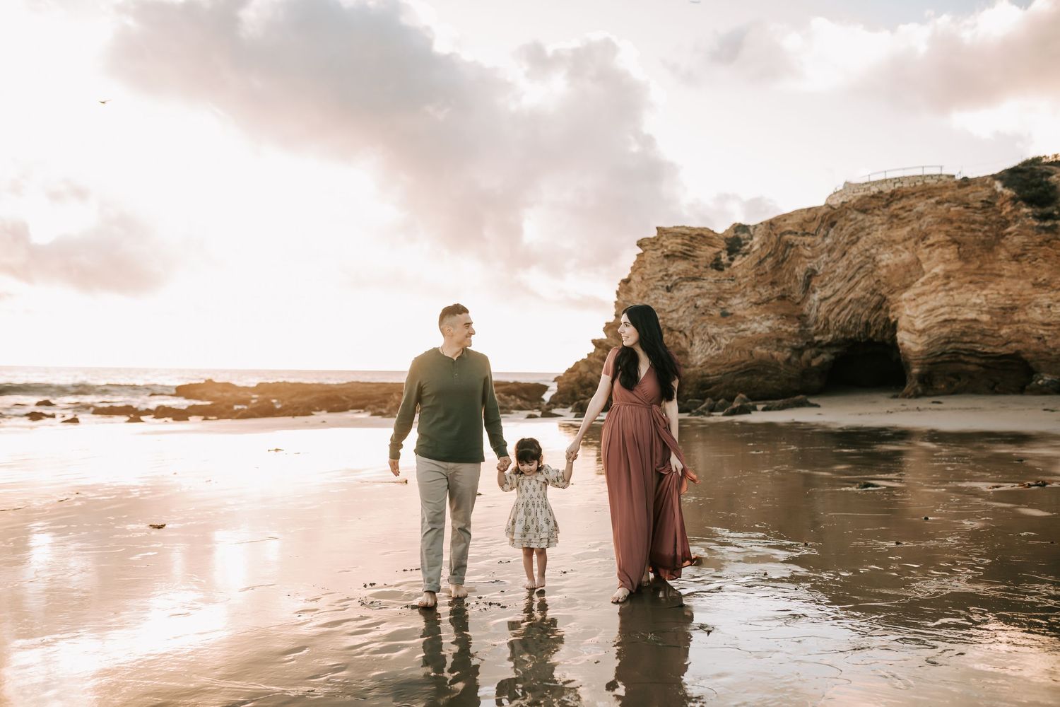 Family silhouettes walk along scenic beach at sunset with dramatic cliffs and coastal scenery in background.