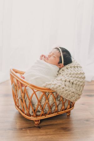Sleeping newborn baby nestled in a cozy woven rattan basket with white blanket.