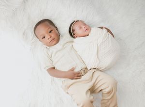 Two young children lying together on white fluffy blanket wearing cream colored outfits.