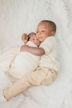 Two siblings cuddling together on soft white fur background in coordinating neutral outfits.
