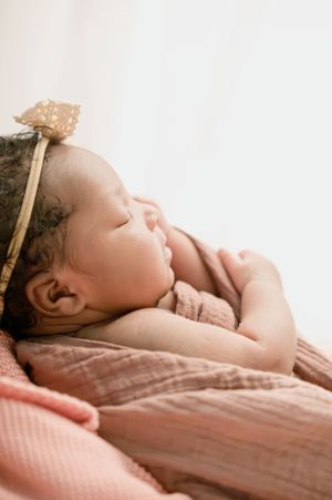 Sleeping baby wearing delicate bow headband resting on pink textured fabric.