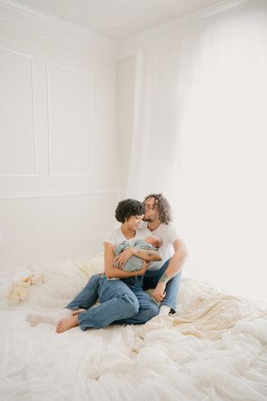 A couple embraces while sitting on cream-colored bedding in a bright room.