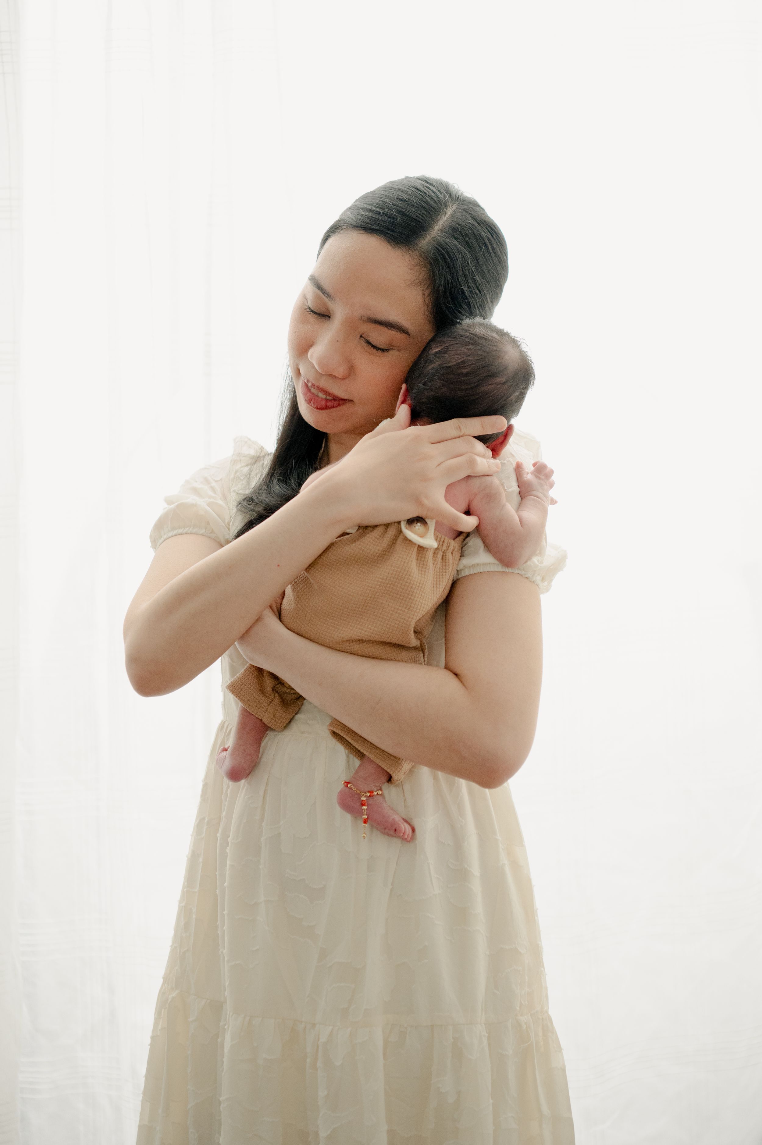 Emotional moment between mother and newborn baby in cream-colored dress against white backdrop.