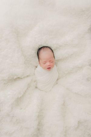 Sleeping newborn baby wrapped in white blanket on soft white textured background.