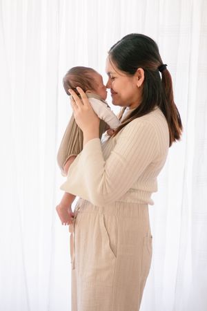 Parent in beige outfit holding newborn baby tenderly against white curtain background.