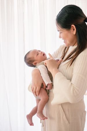 Mother in beige sweater holding and smiling at baby near white curtains.