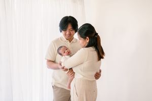 Family embracing while holding newborn baby in minimalist white room setting.