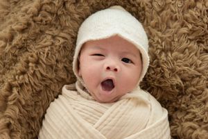 Infant wearing white cap yawning while lying on textured brown fabric.