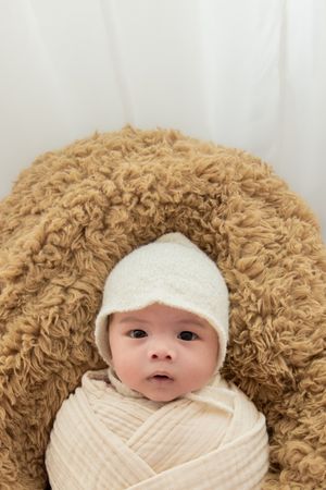 Baby wrapped in cream fabric with white cap against brown textured background.