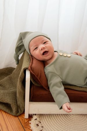 Baby in sage green outfit and white hat lying on wooden surface with textured blanket.