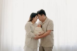 Parents in neutral colors holding newborn baby against bright window backdrop.