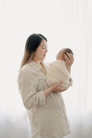Mother in striped shirt holding and gazing lovingly at newborn against white backdrop.