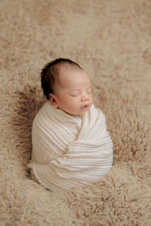 Sleeping newborn baby wrapped in soft white fabric against a beige textured background.