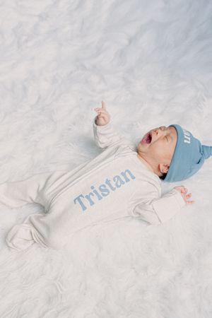 Baby in white outfit and blue hat lying on white blanket.
