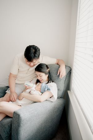Family sharing intimate feeding moment on grey couch by window.