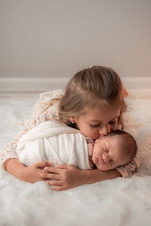 A child gently embraces a sleeping baby on soft white bedding.