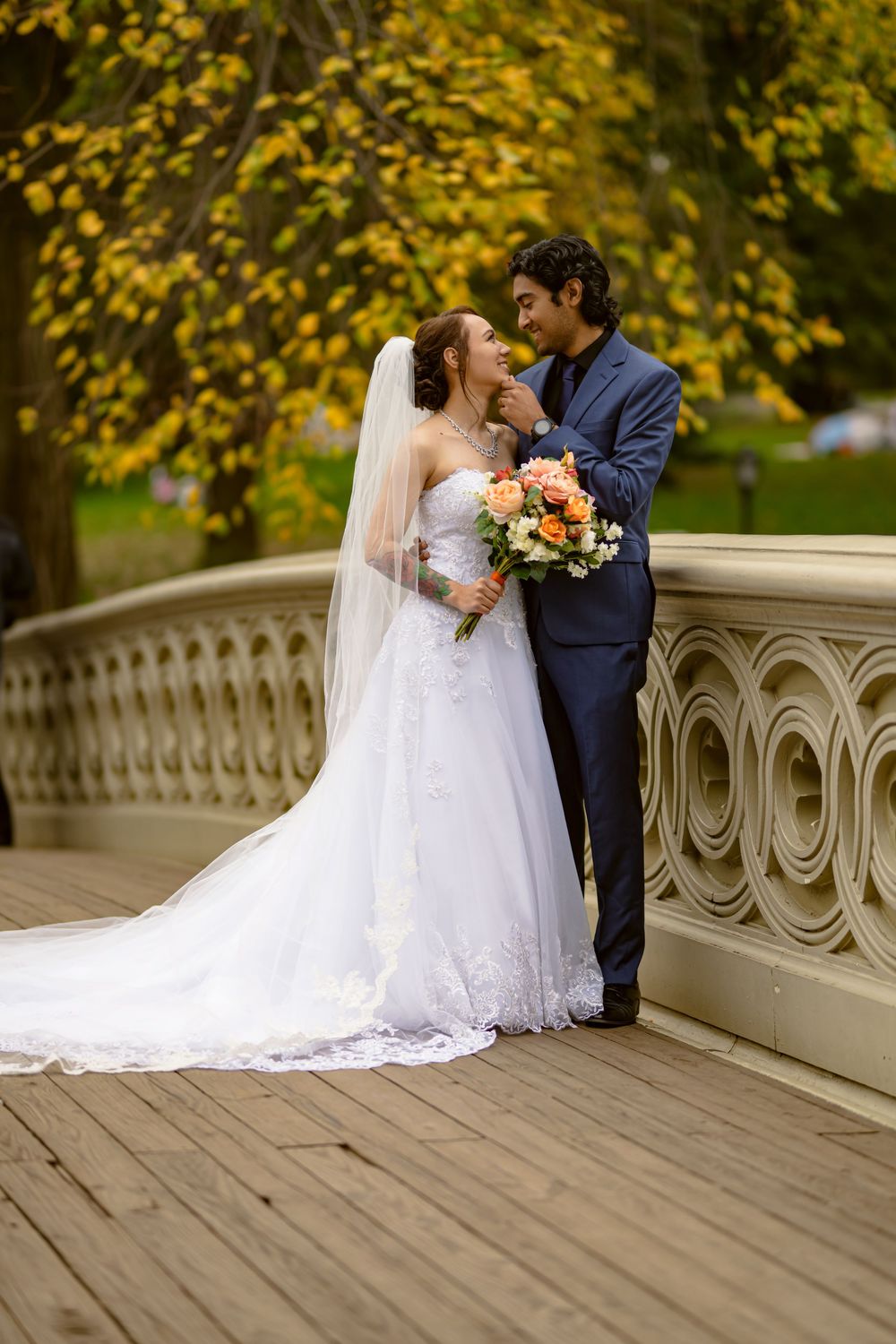 Elegant wedding couple sharing intimate moment on decorative bridge with autumn trees in background.