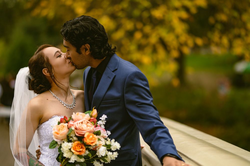Bride in lace gown holds bouquet while posing with groom against autumn scenery.
