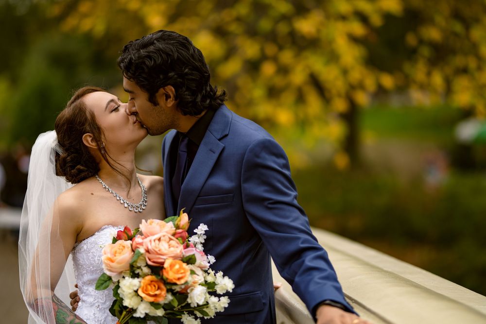 Wedding couple poses on ornate bridge with golden autumn leaves creating romantic backdrop.