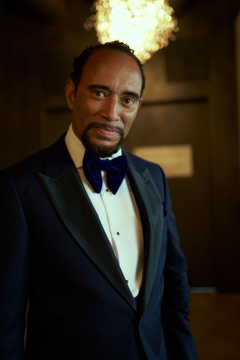 Portrait of a person in navy tuxedo and bow tie illuminated by warm overhead lighting. The Bordone LIC