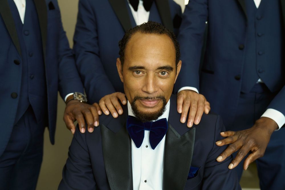 A close-up shot of a person in a black tuxedo and bow tie with hands on their shoulders. The Bordone LIC