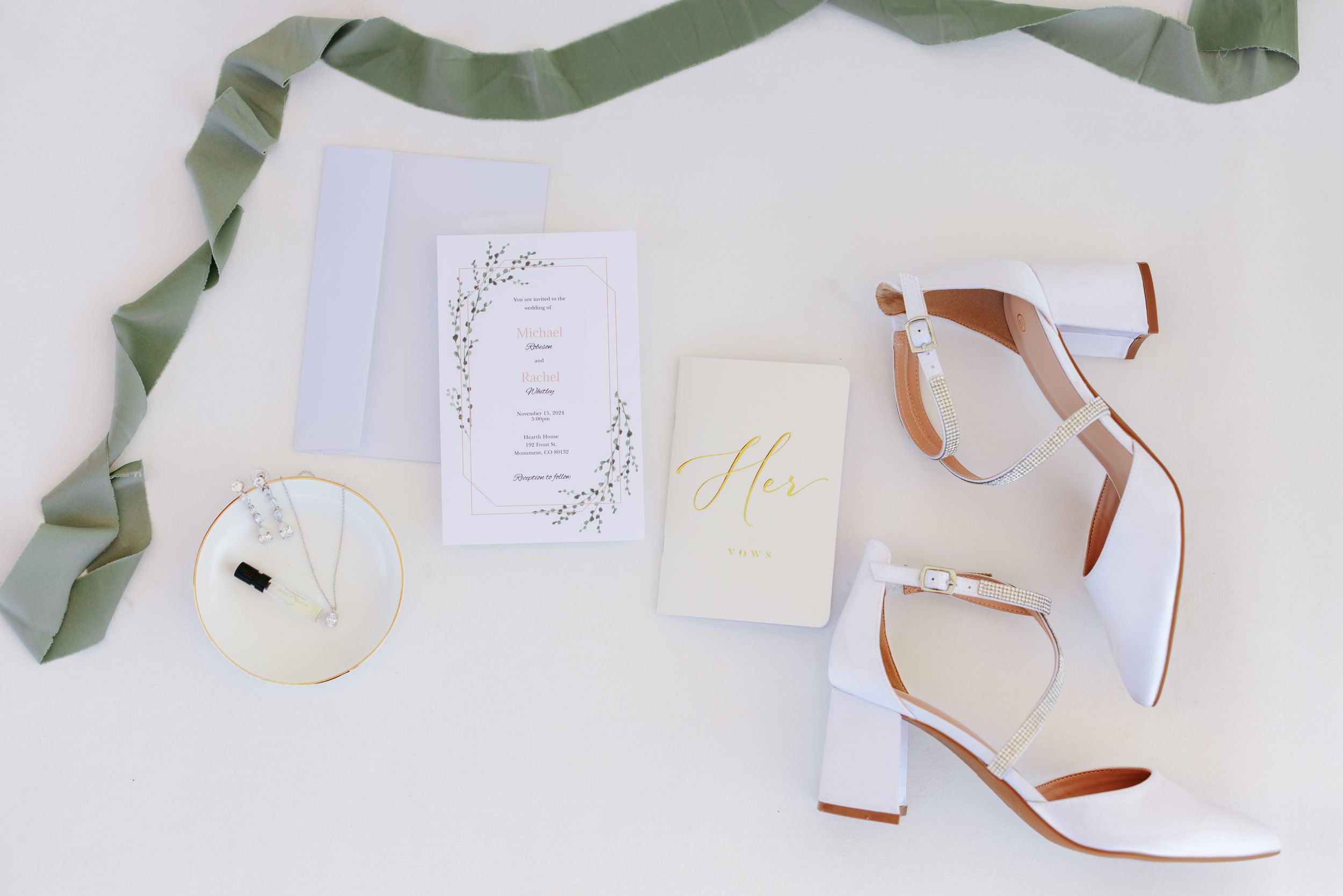 Wedding details flatlay with white heels, invitation suite, and sage green ribbon on light background.