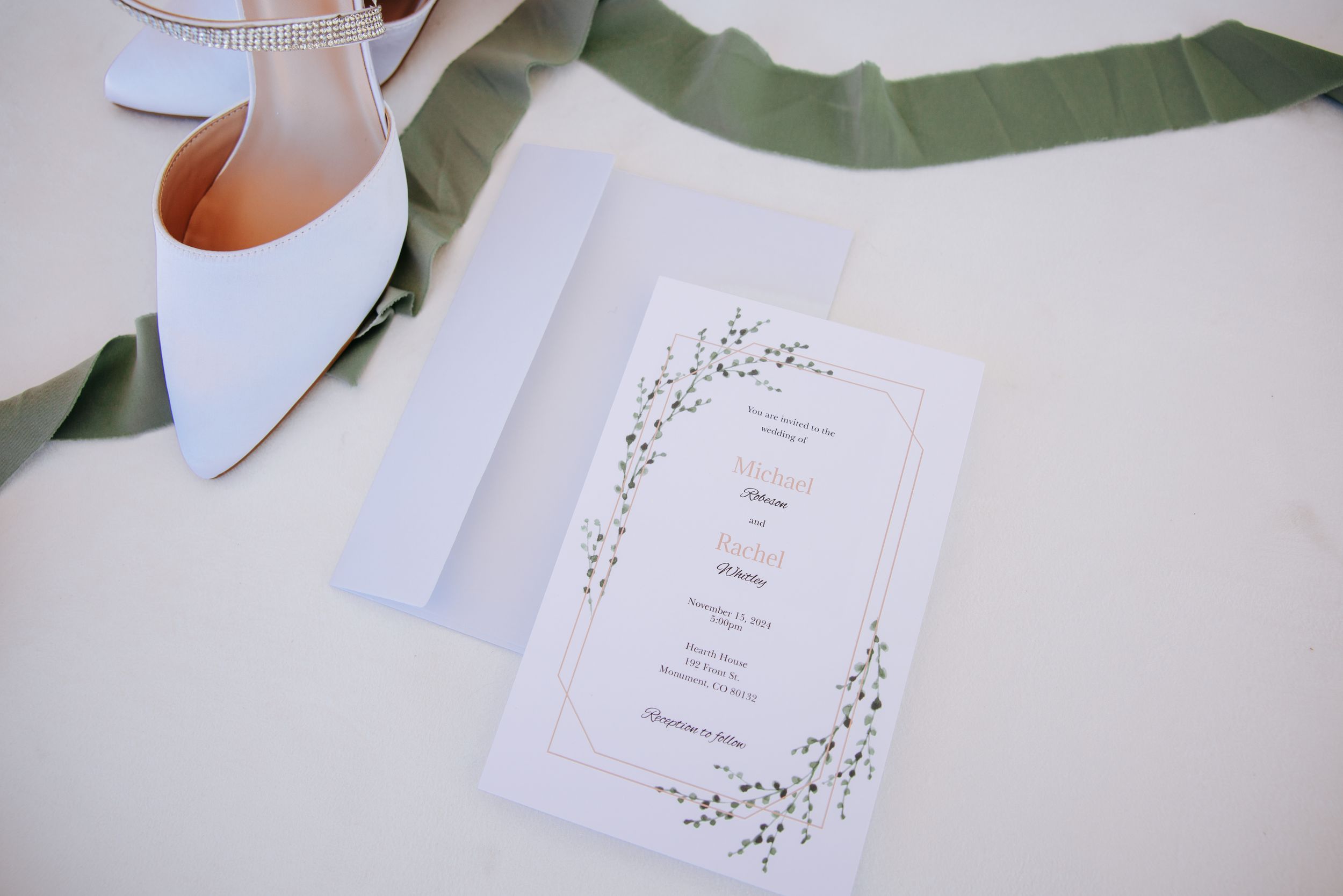 Light blue wedding shoe next to an elegant white wedding invitation with delicate floral design on white surface.