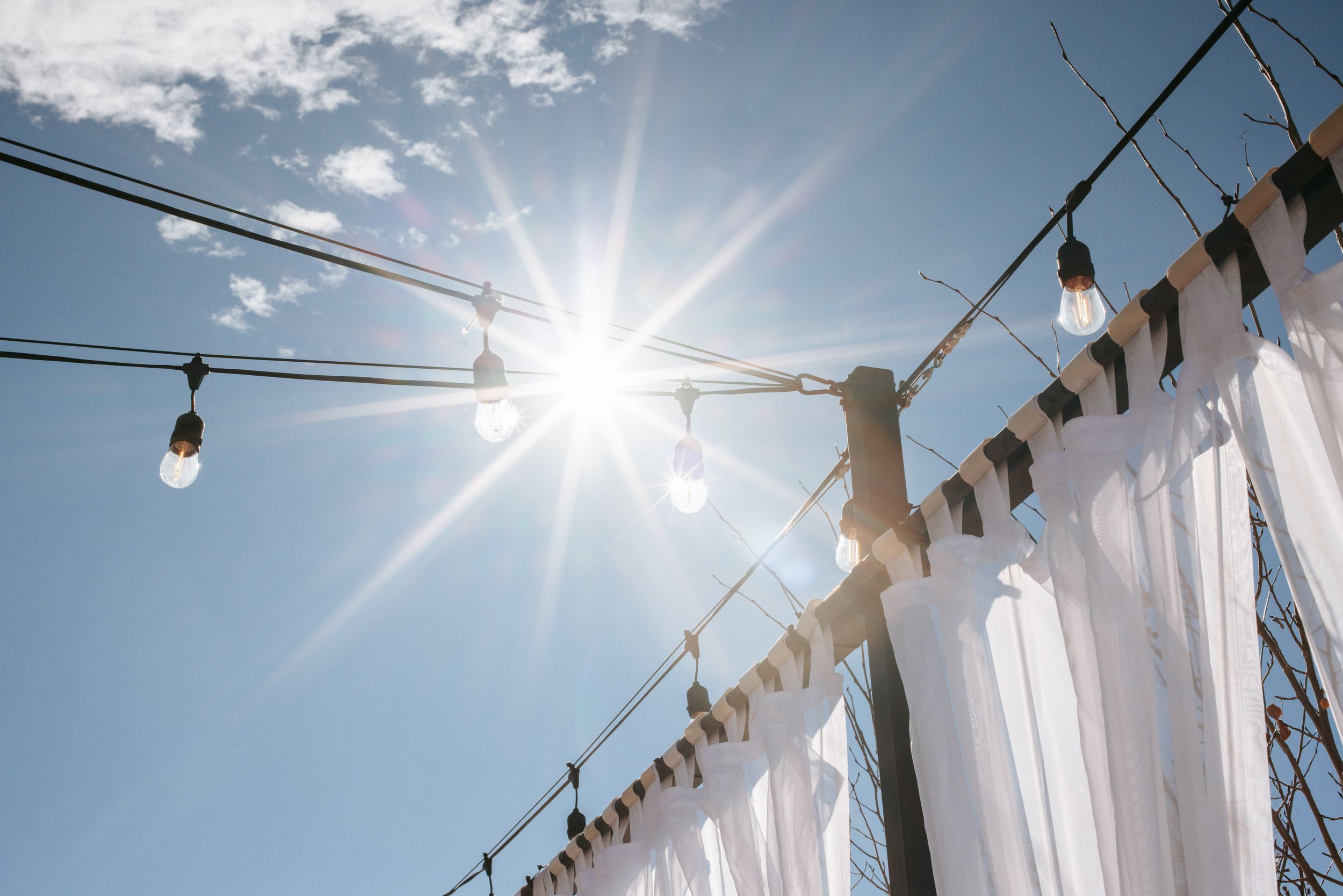 String lights hang against a bright blue sky with white curtains and sunlight creating a romantic outdoor setting.