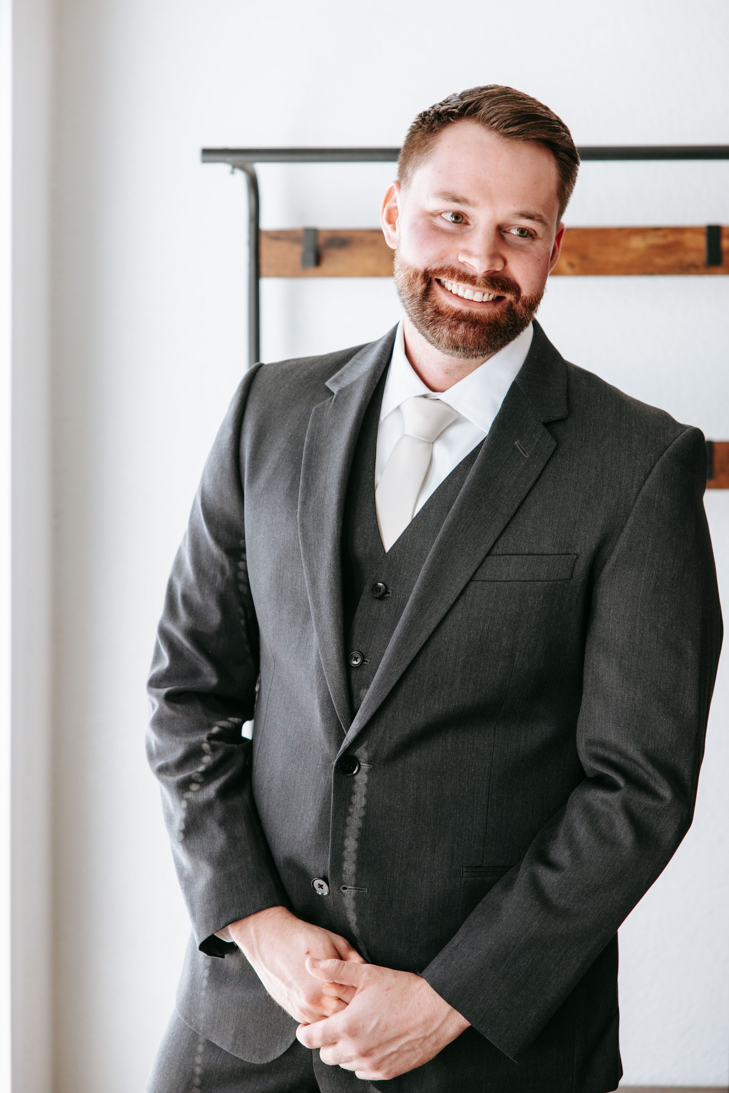 A person wearing a dark gray three-piece suit stands in a bright room with rustic decor.