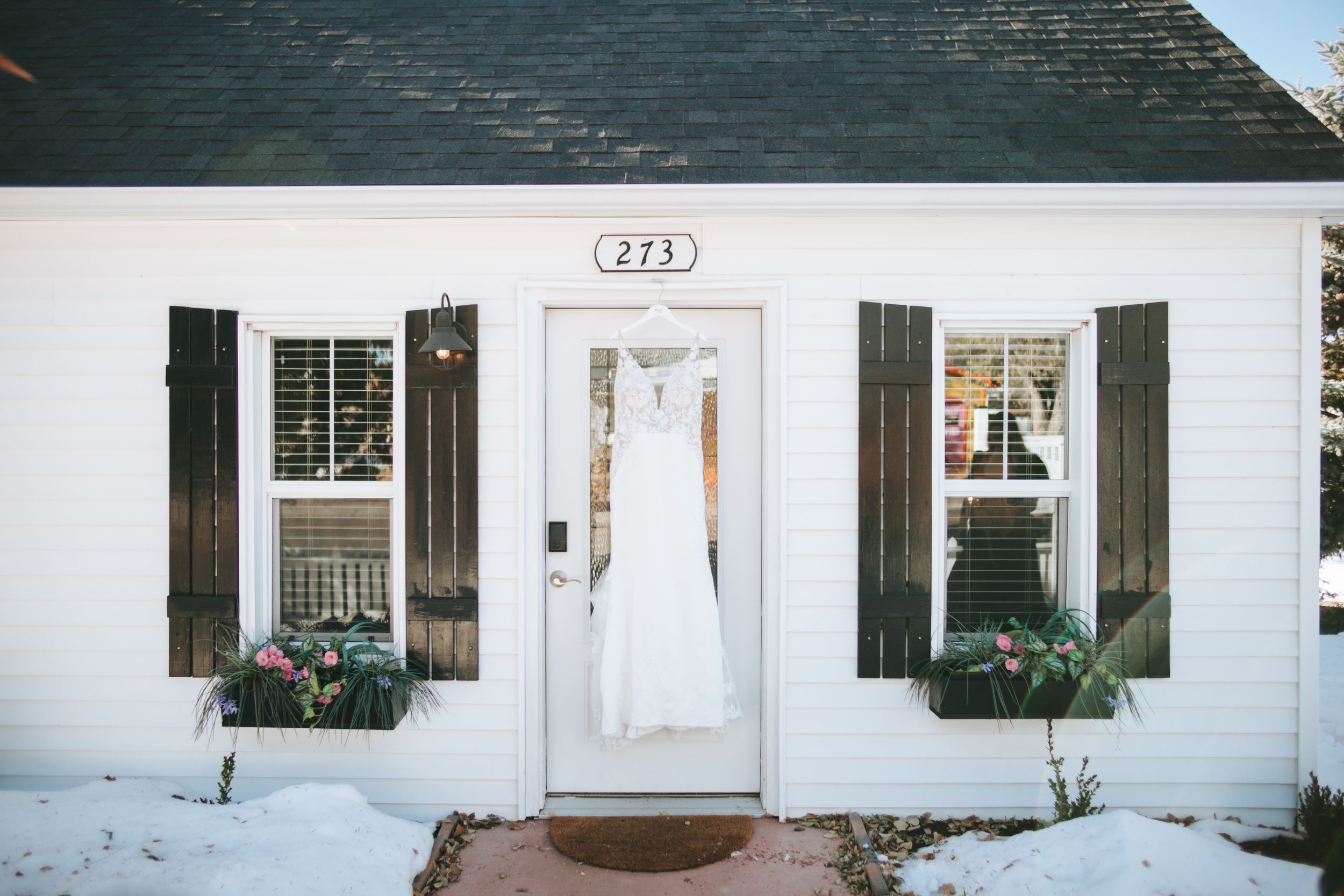 Wedding dress displayed in window of white house with black shutters and flower boxes in winter setting.
