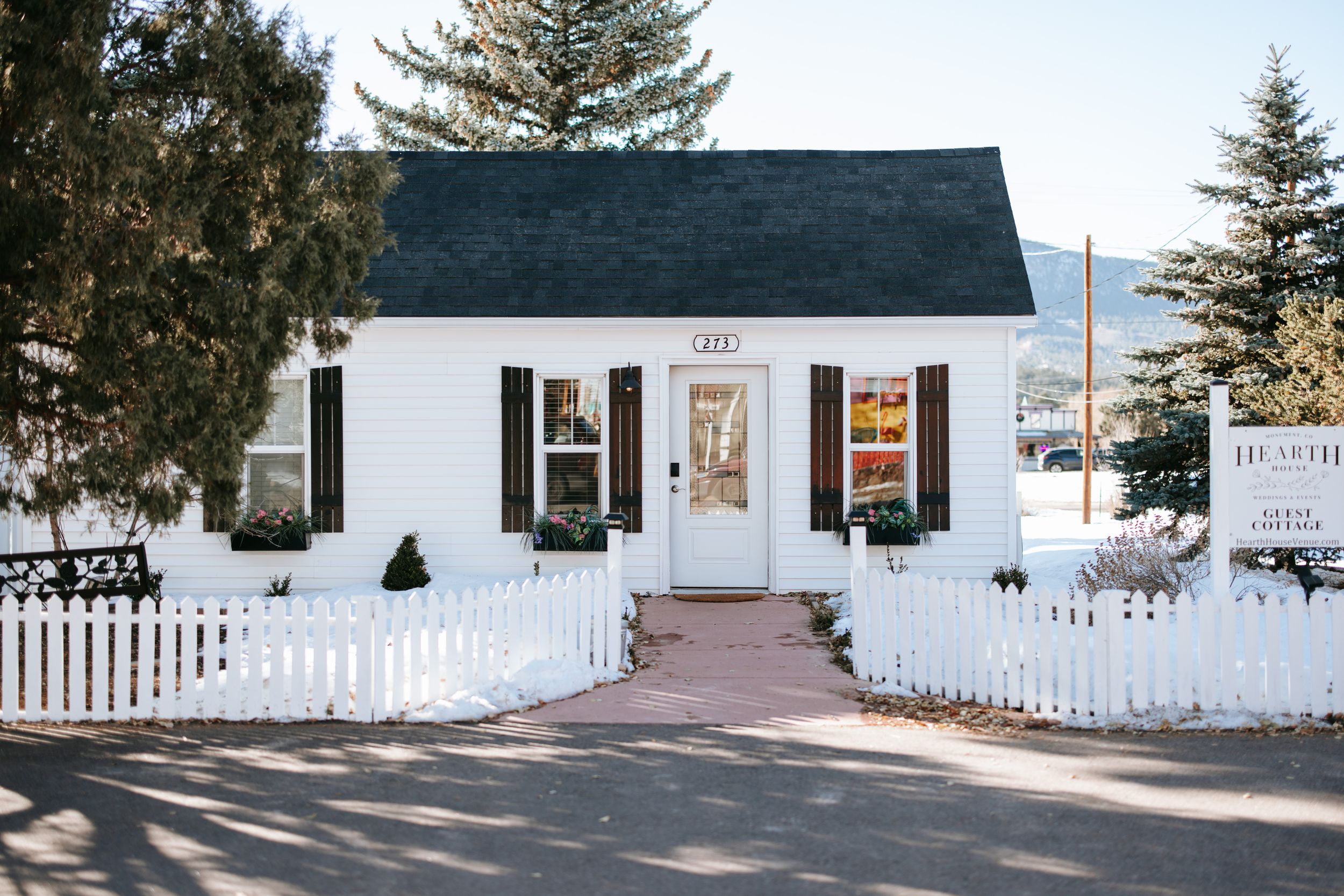 Charming white cottage-style house with black shutters and white picket fence surrounded by snow and evergreen trees.