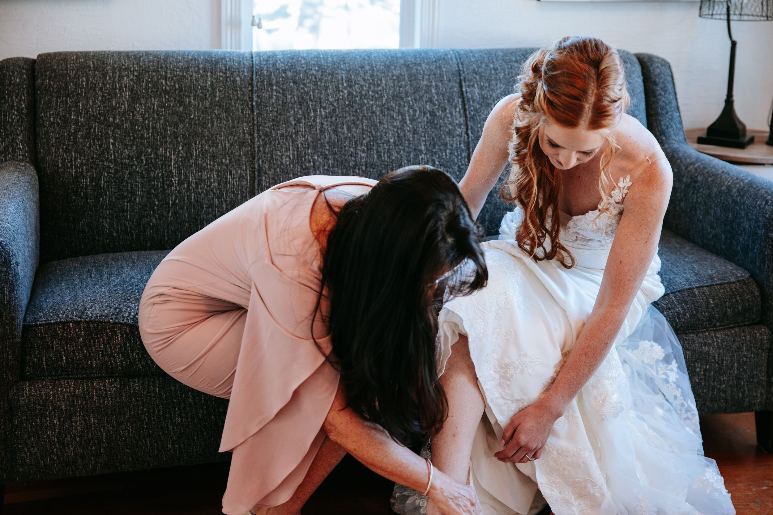 A woman in a wedding dress sitting on a dark grey couch getting help with shoes.