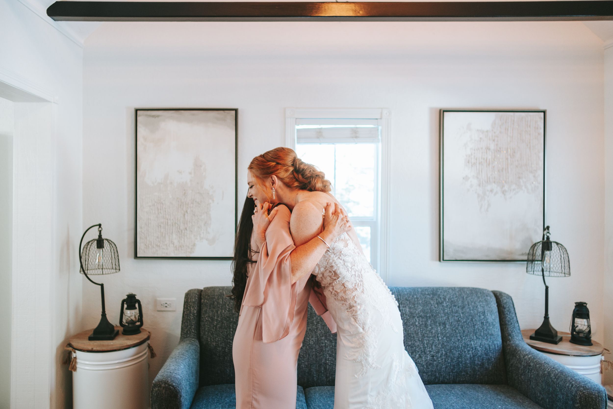 Two figures embrace in an indoor setting with white walls, framed art prints, and matching decor accents.
