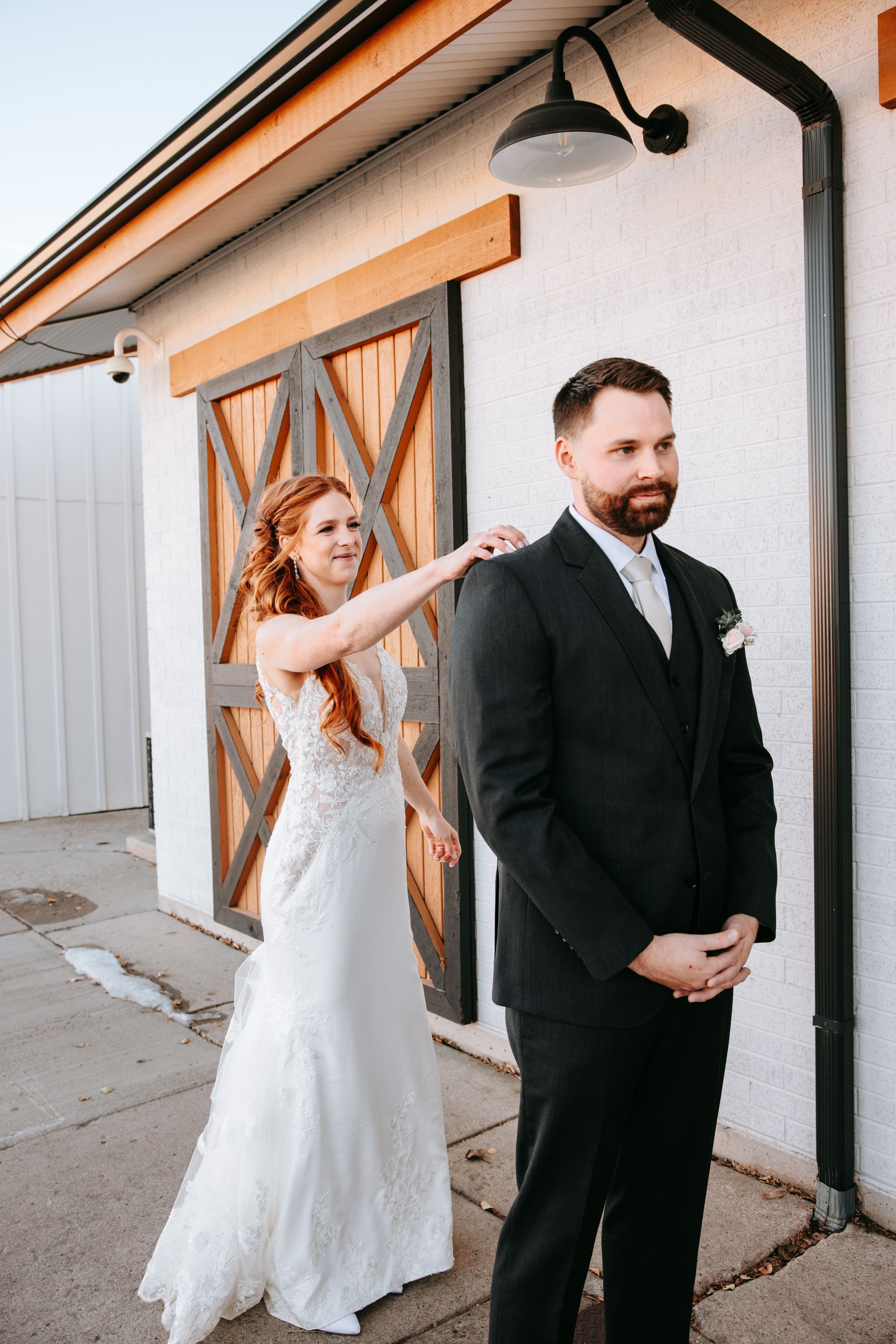A couple enjoys a sweet first look moment before their wedding ceremony outside a white building with wooden barn doors.