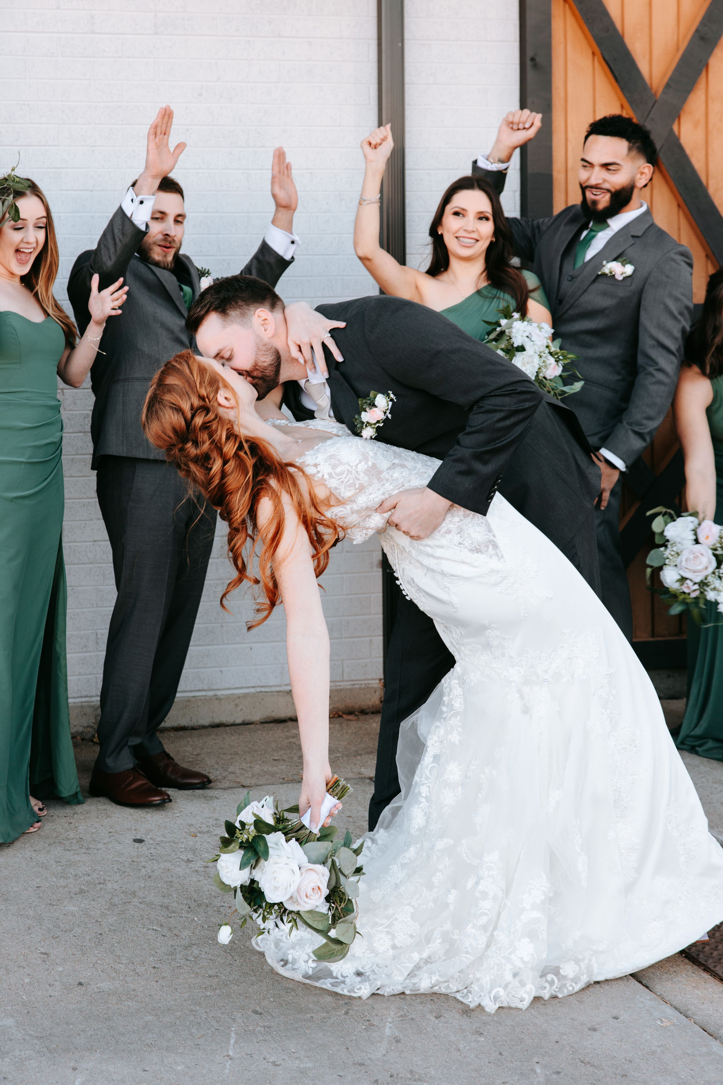 Wedding party poses playfully outside with white flowers and greenery while wearing dark suits and emerald dresses.