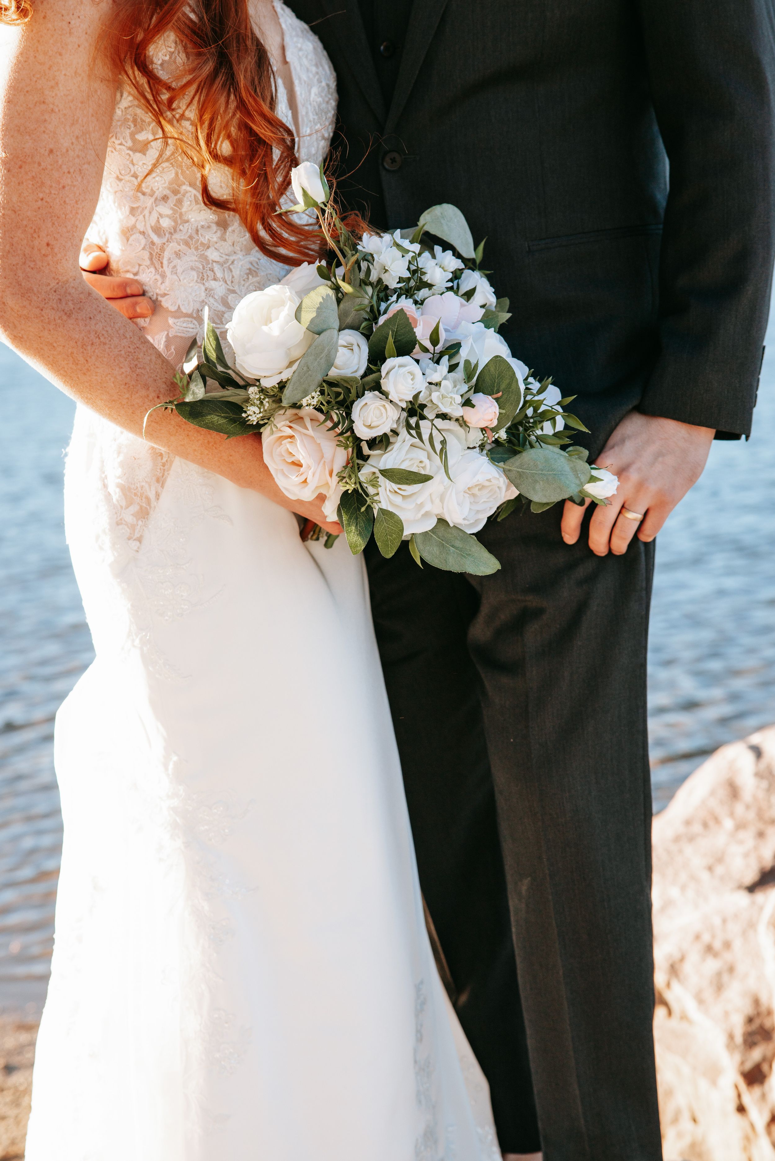 A close-up of a wedding bouquet with white roses and greenery held between a white dress and black suit.