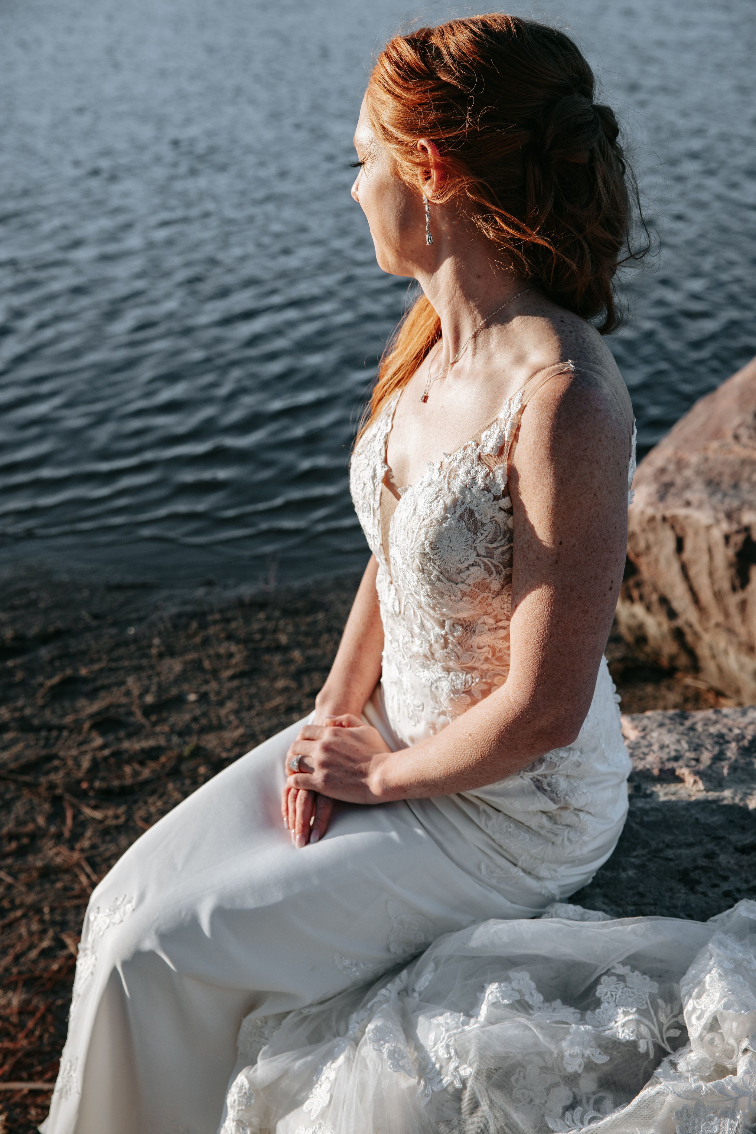 A bride in a white lace dress sits gracefully by the water's edge during sunset.