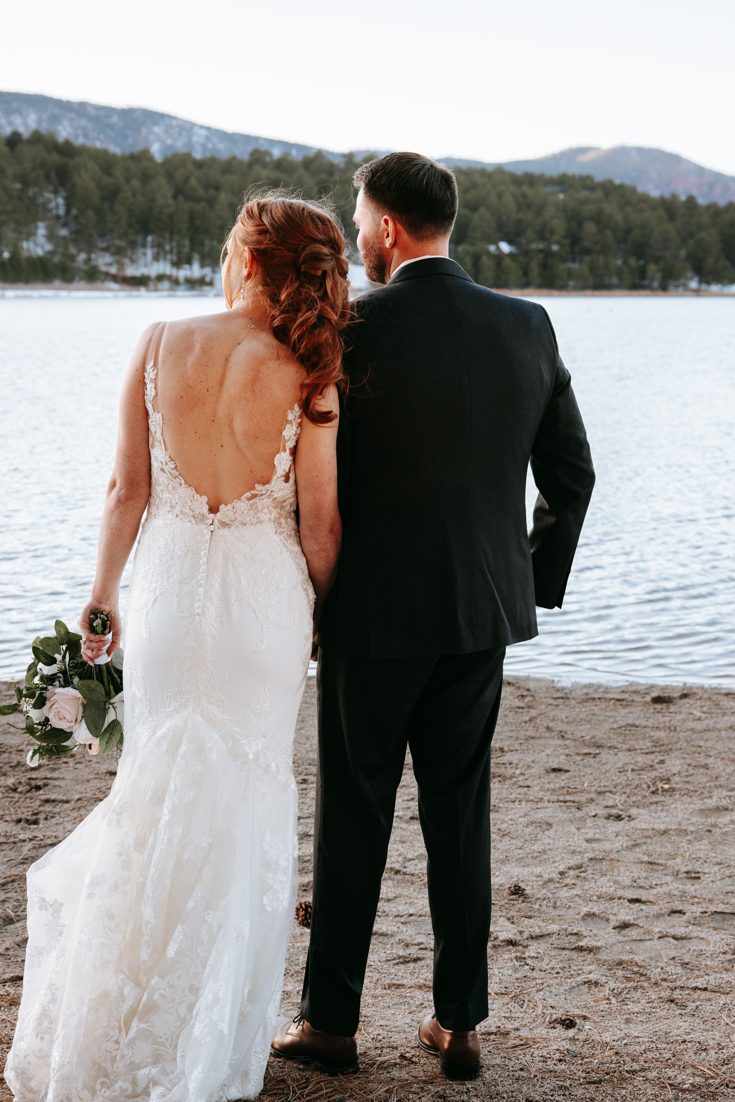 A wedding couple stands together at a snowy lakeside with mountains in the background during their winter celebration.