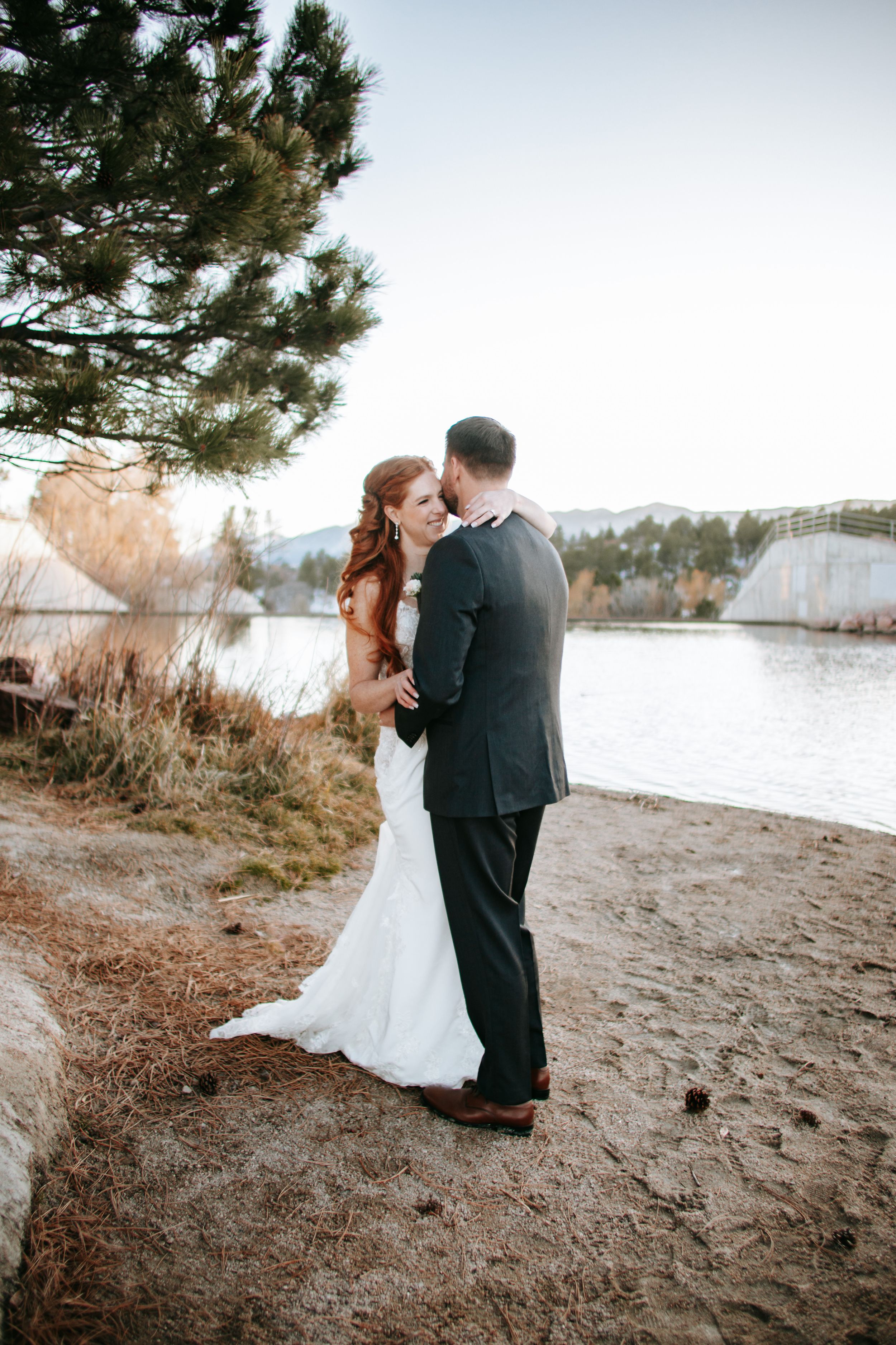 A romantic wedding couple embraces on a lakeside beach surrounded by pine trees at golden hour.