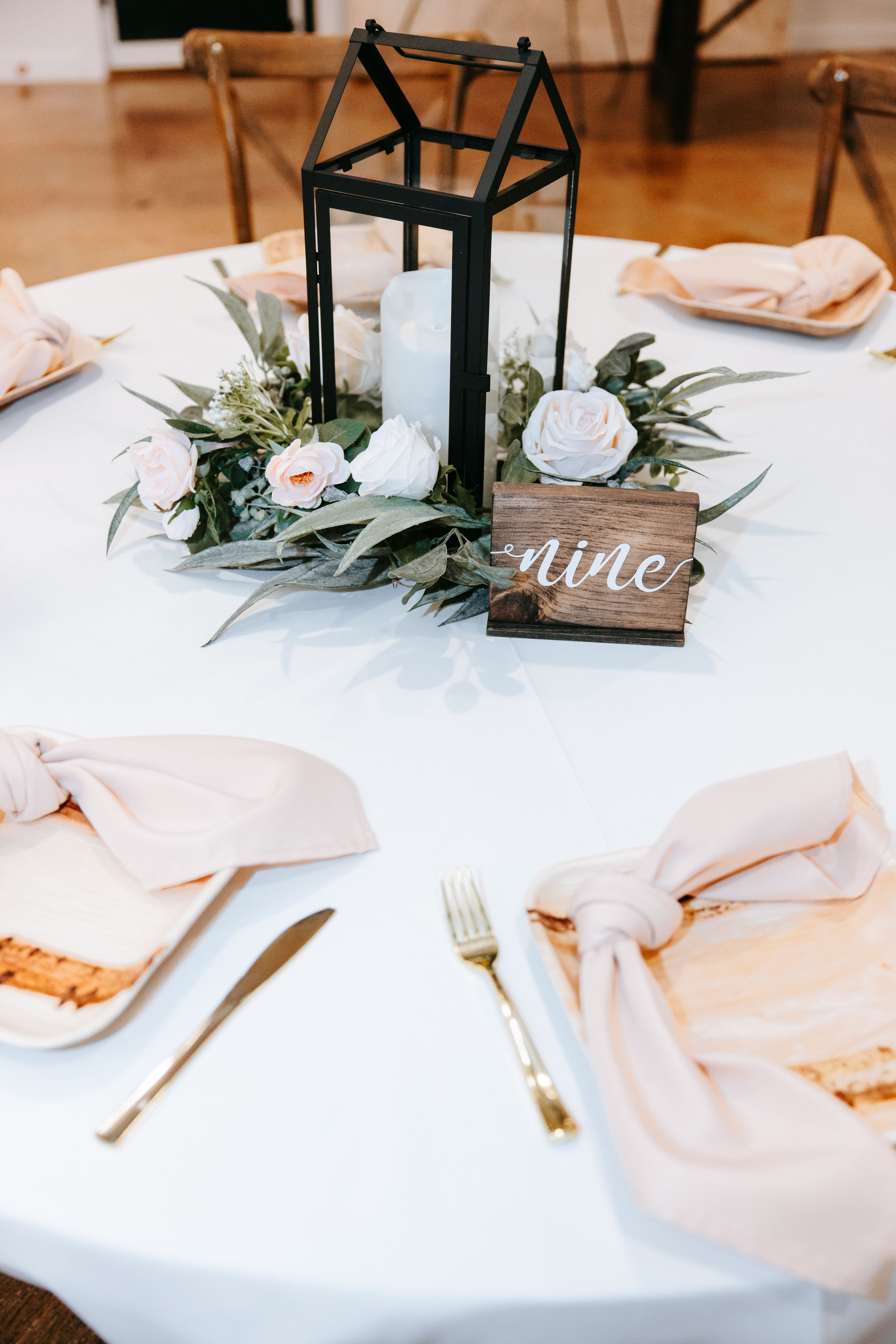Wedding reception table centerpiece with black lantern, white candle, pink roses and greenery on white tablecloth.