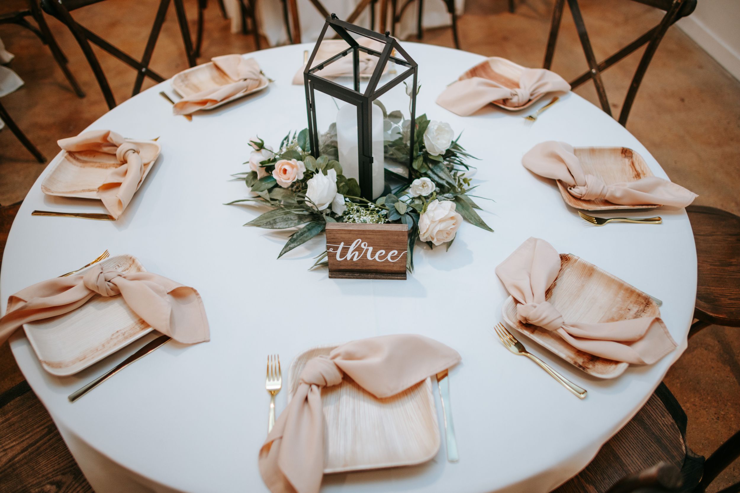 Elegant wedding table setting with black lantern centerpiece, blush napkins, and fresh greenery on white tablecloth.