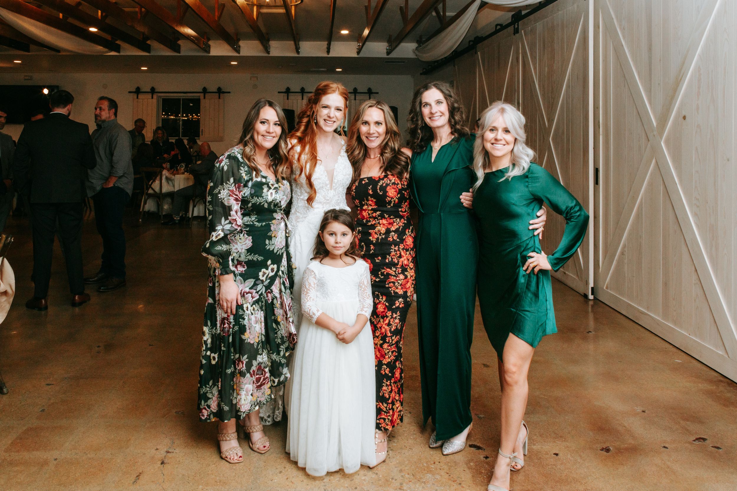 Women in elegant floral and jewel-toned dresses pose together at an indoor event space with wooden floors and white walls.