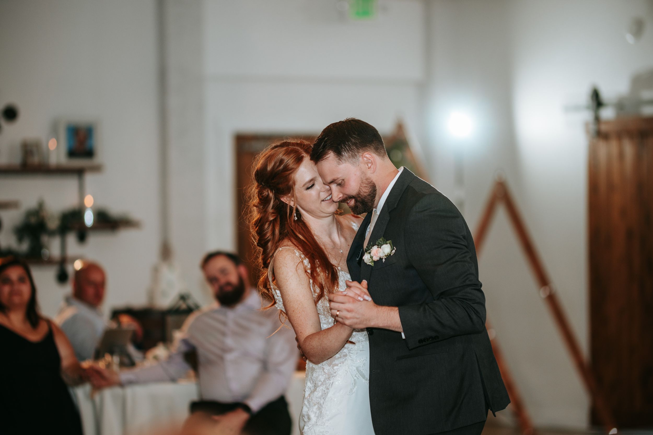 A couple shares their first dance at a wedding reception in a rustic indoor venue with guests watching from the side.