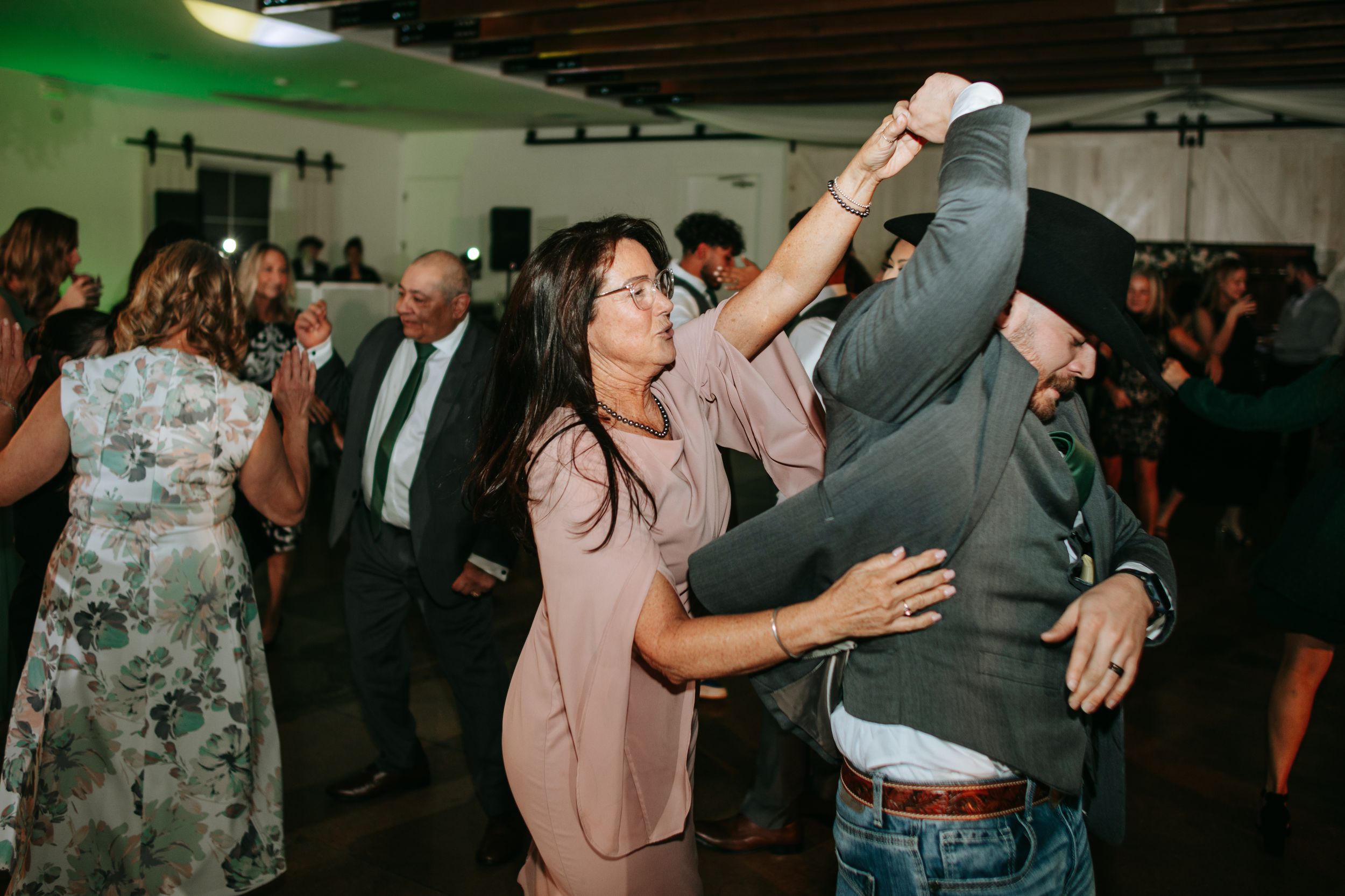 A lively crowd dances and celebrates at a reception venue with warm lighting and wooden beams overhead.