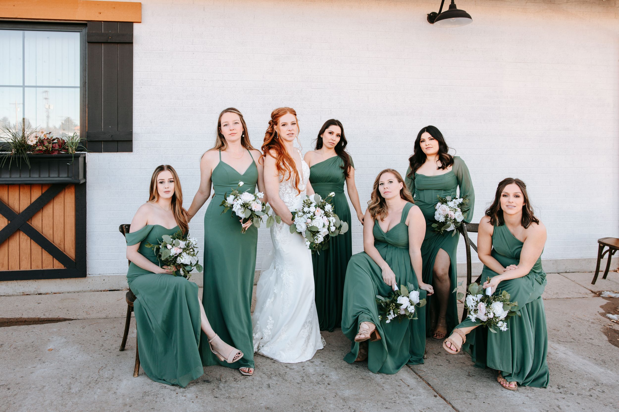 Wedding party in emerald green bridesmaid dresses posing with bride in white gown against white building exterior.