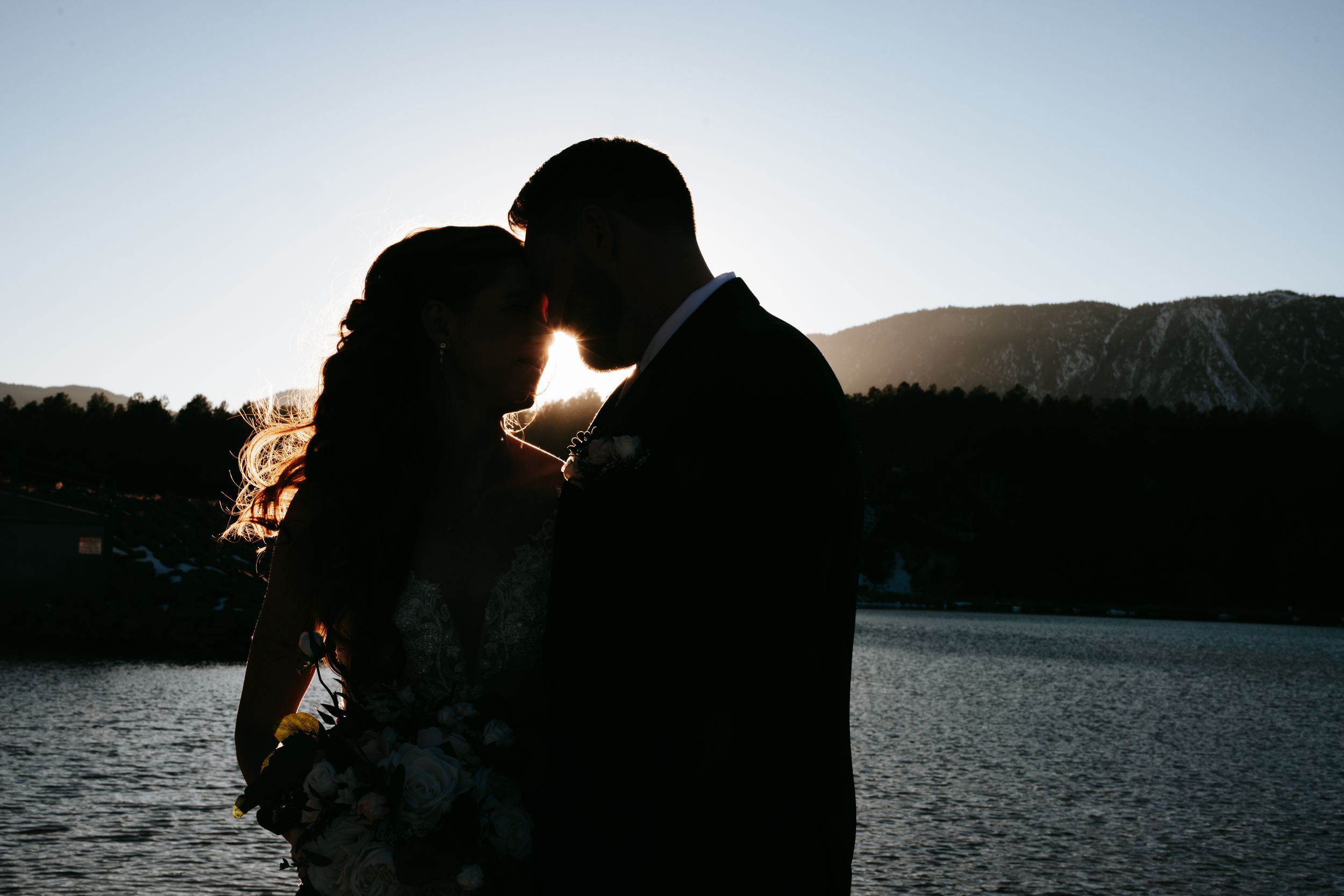 Sunset silhouettes of two figures embracing by a tranquil lake, creating a romantic golden hour moment.