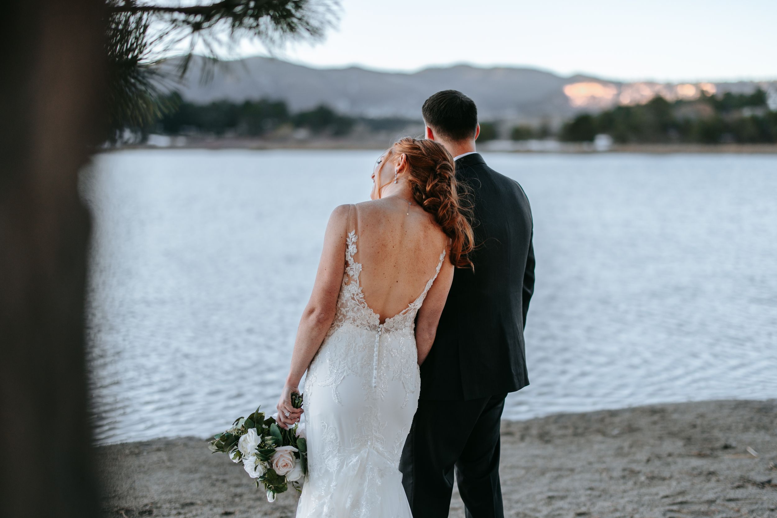 A wedding couple stands close together by a lakeside at sunset with mountains in the background.